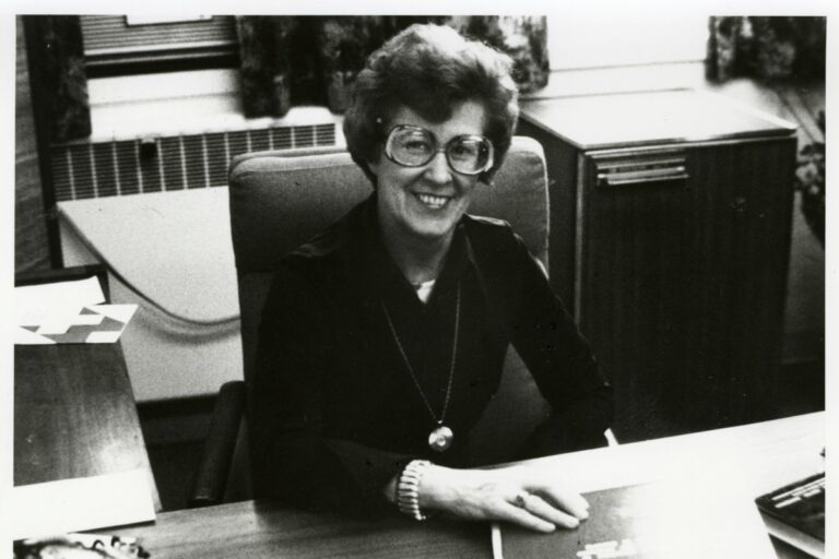 A woman in glasses, Loretta Ford, sits at a desk surrounded by papers, reflecting on her time as a former nursing dean.