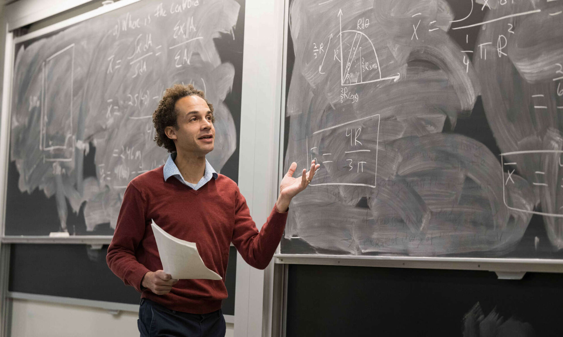 Mark Buckley holds papers and gestures at a chalkboard full of equations during a biomechanics class.