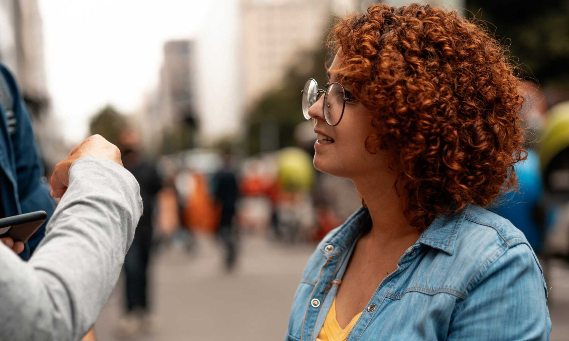 Woman with large oversized glasses looks on and listens as two people nearby off-camera speak and gesticulate.