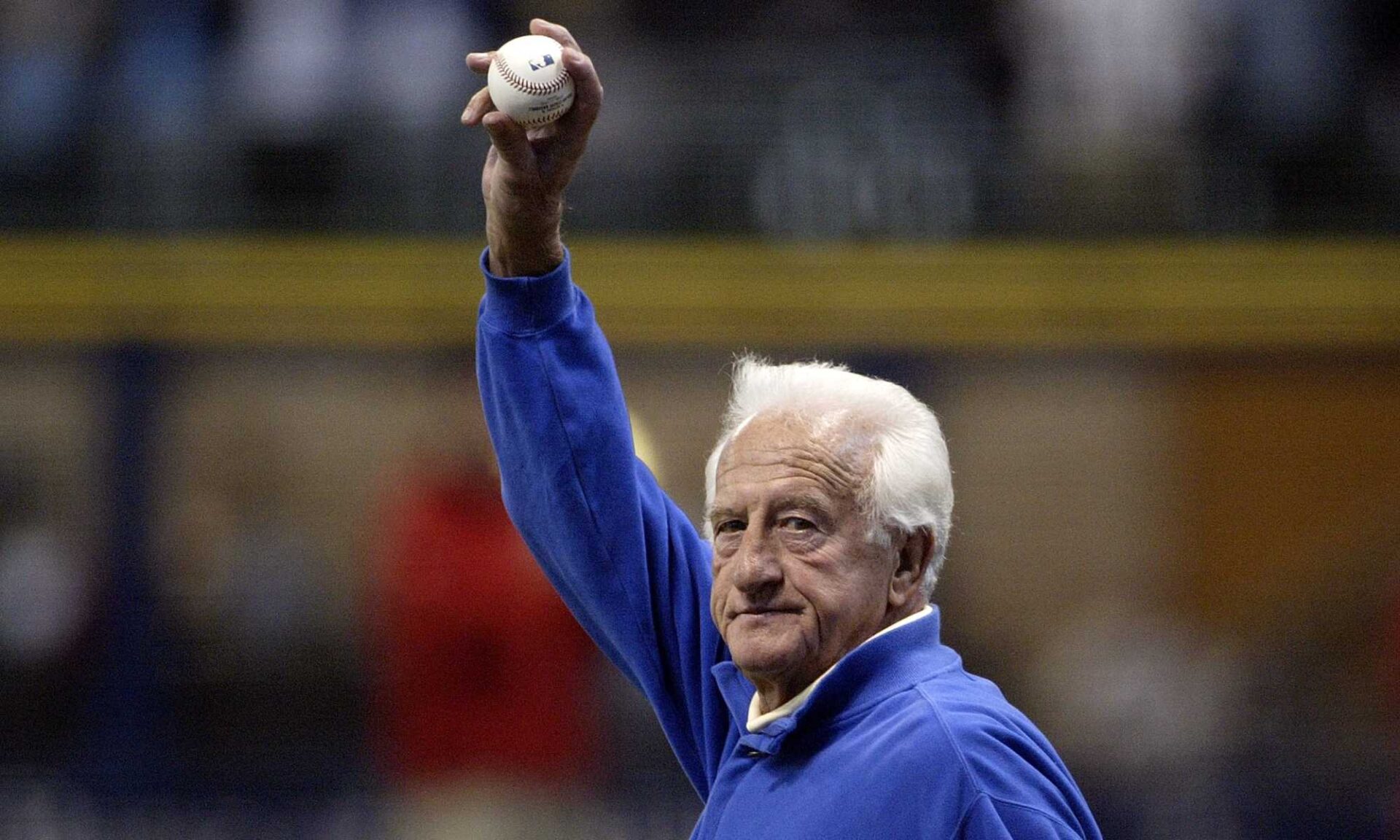 Bob Uecker in a blue zip-up holds a baseball over his head in a crowded stadium.