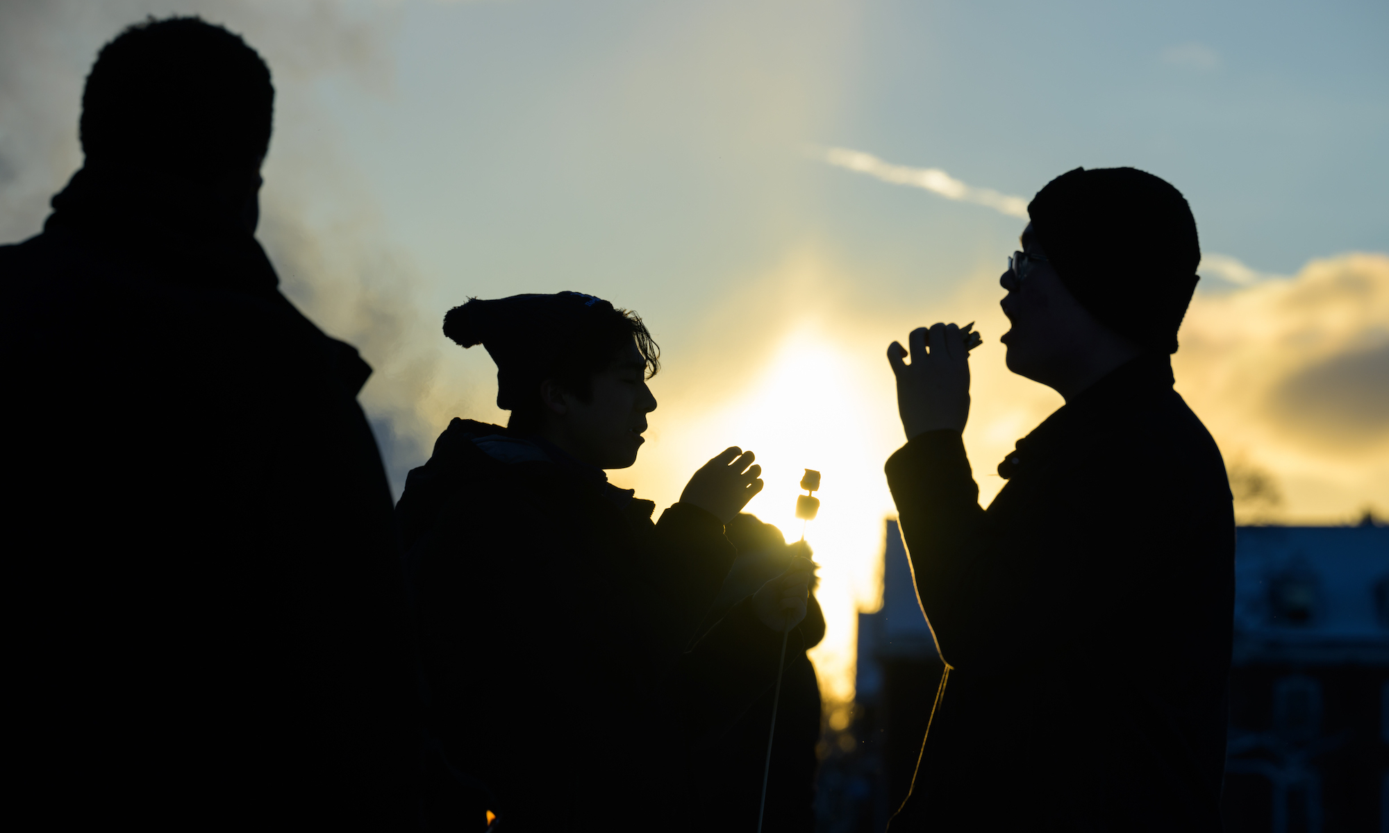 Three students in shadow enjoying s'mores as the sun sets.