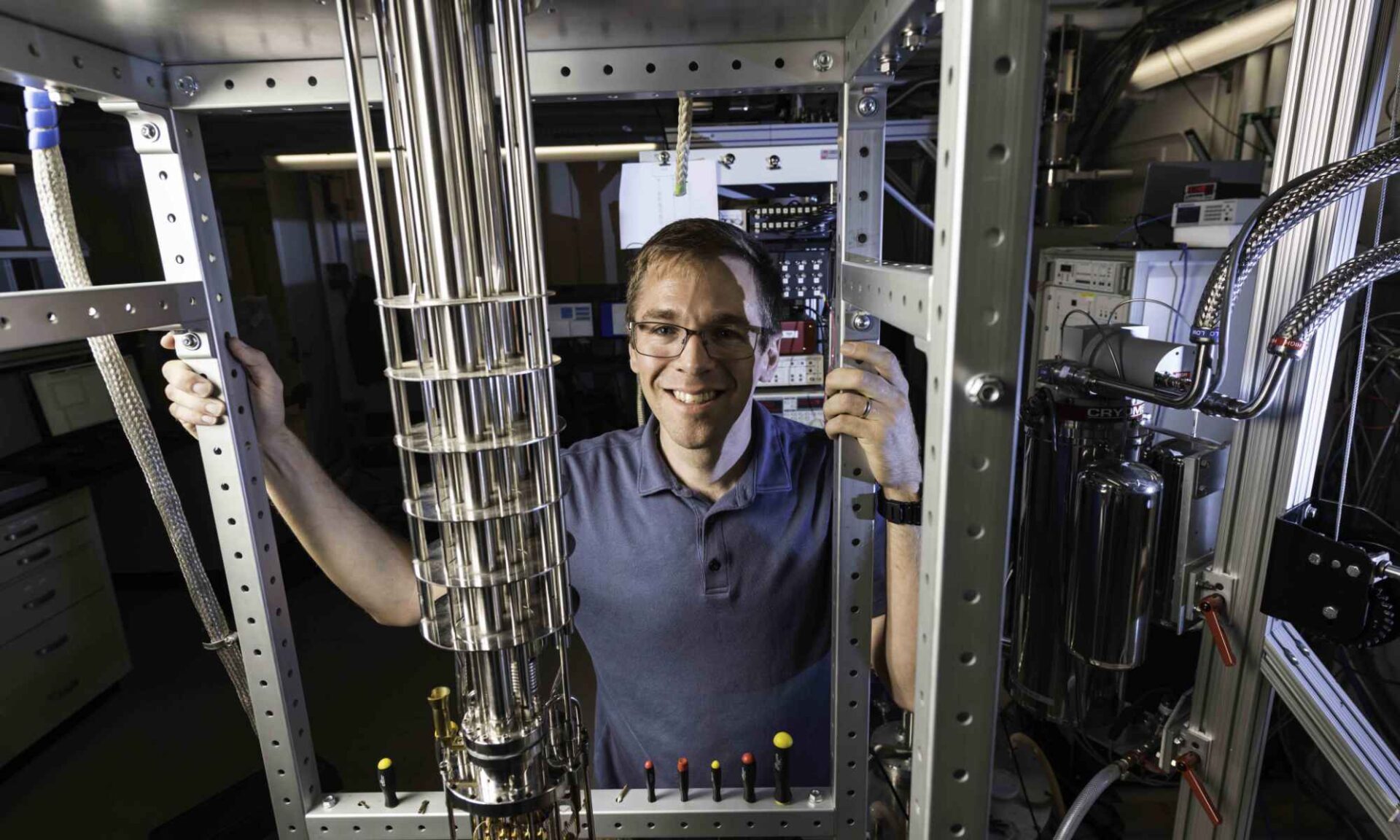 John Nichol standin behind a dilution refrigerator, used to study quantum systems, including nuclear-spin dark states.