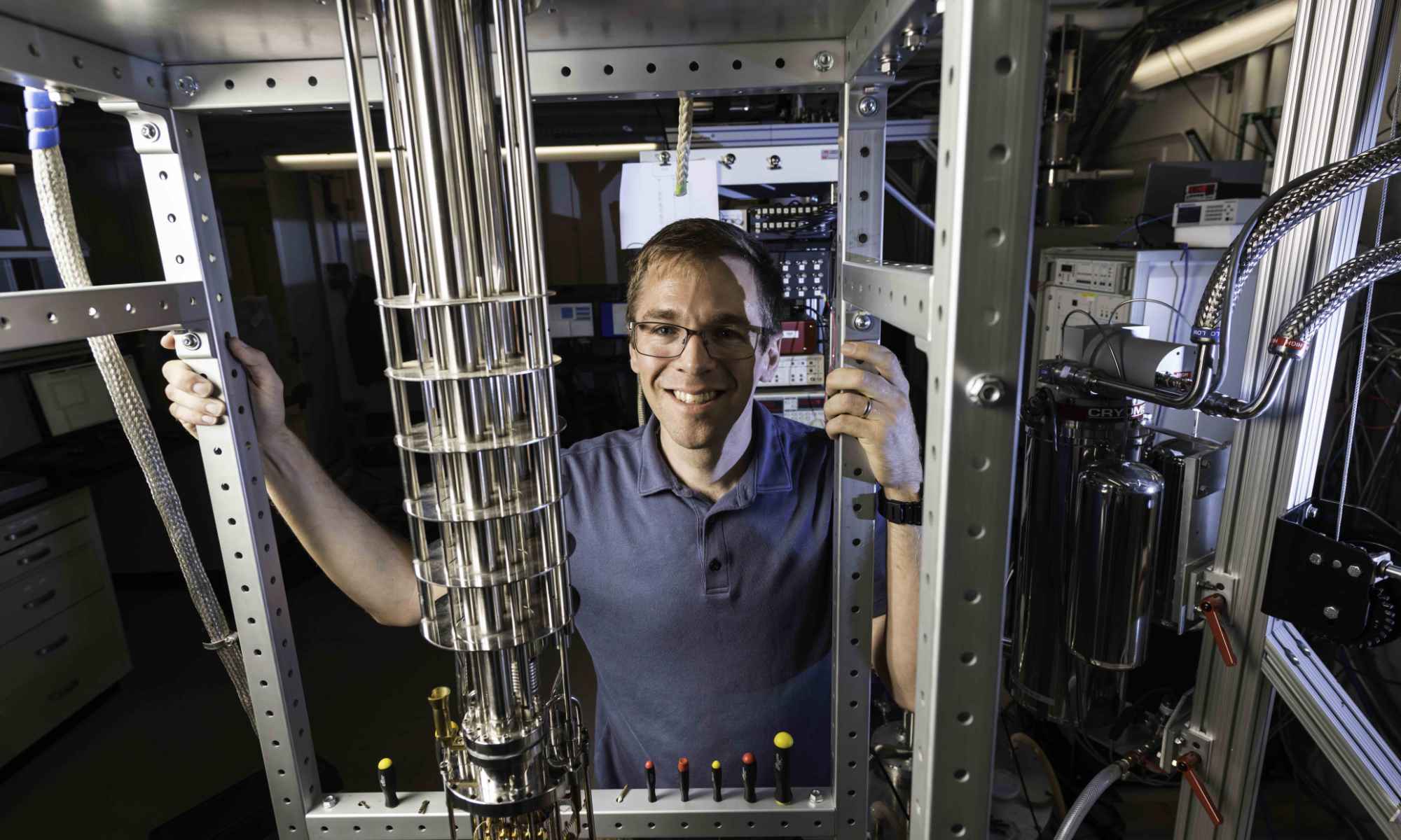 Feature image John Nichol standin behind a dilution refrigerator, used to study quantum systems, including nuclear-spin dark states.