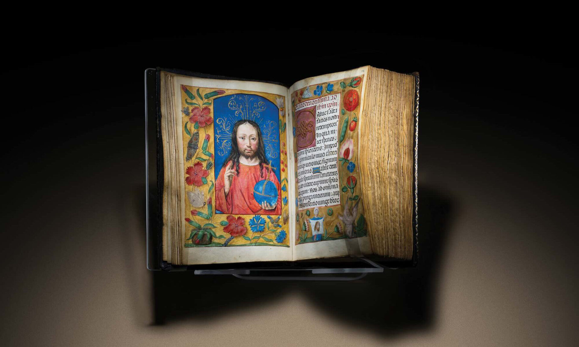 The Ghent-Bruges Book of Hours sits open to colorfully painted spread with a portrait of a saint opposite calligraphic text, both surrounded by flowers and plants.