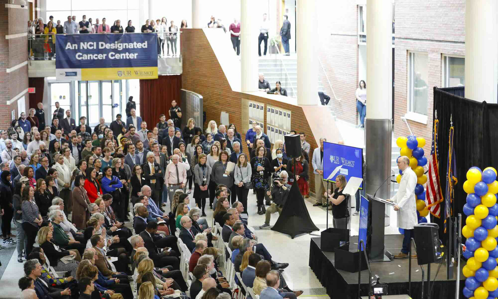 Jonathan Friedberg addresses a large crowd at Wilmot Cancer Institute, with a banner reading 'An NCI Designated Cancer Center' in the background.