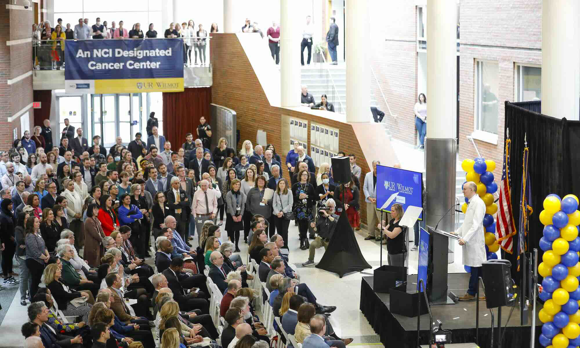 Jonathan Friedberg addresses a large crowd at Wilmot Cancer Institute, with a banner reading 'An NCI Designated Cancer Center' in the background.