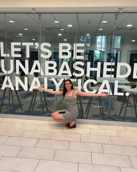 Farrell Cooke crouches down in front of a glass wall with the words 'Let's Be Unabashedly Analytical.'