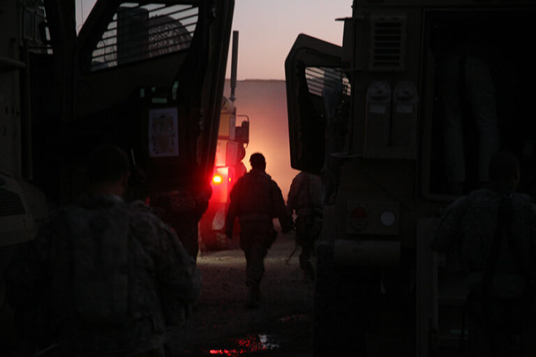A US solder in uniform walks toward the setting sun in between army vehicles and machines, all in silhouette.