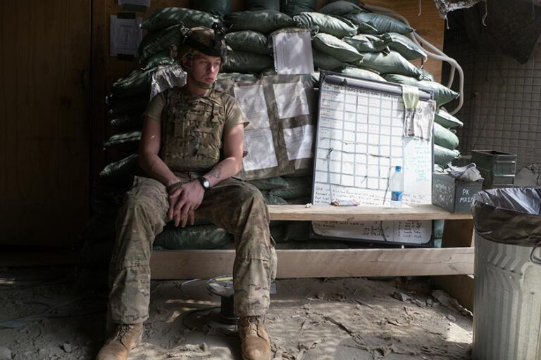 A US soldier in uniform sits and stairs off camera with supplies stacked behind him.