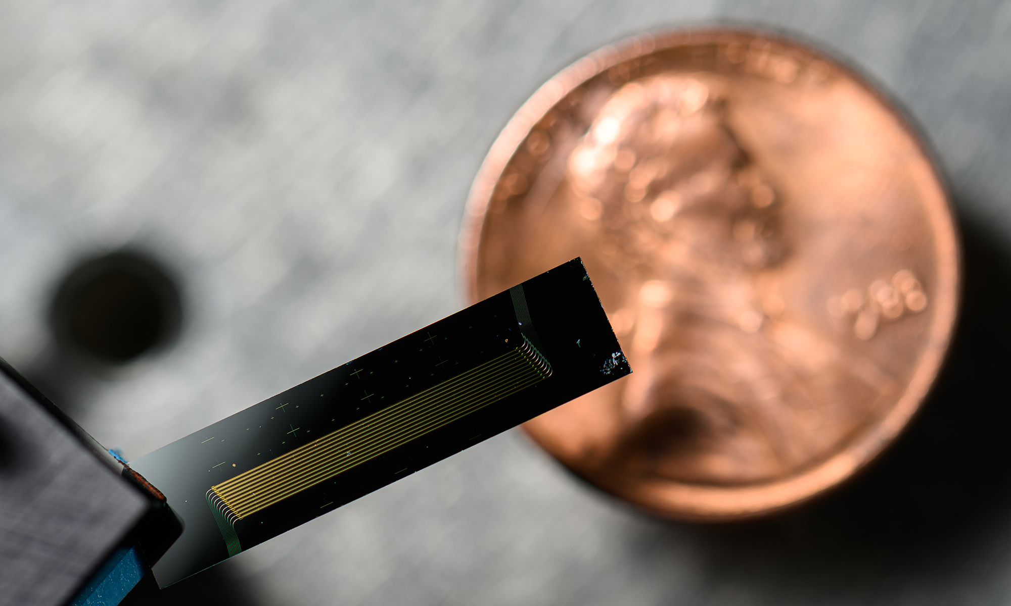 Close-up of a chip-scale laser held in front of a penny, highlighting its miniature size and intricate gold circuitry.