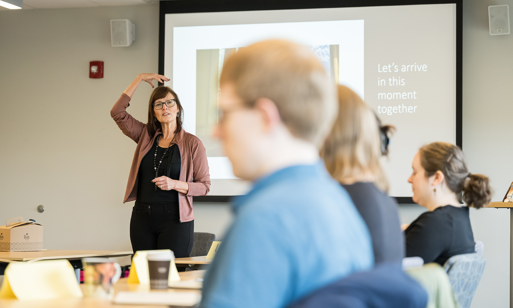 Woman leads a mindfulness exercise in a classroom, gesturing with one hand raised, as students listen with a slide reading 'Let’s arrive in this moment together'.