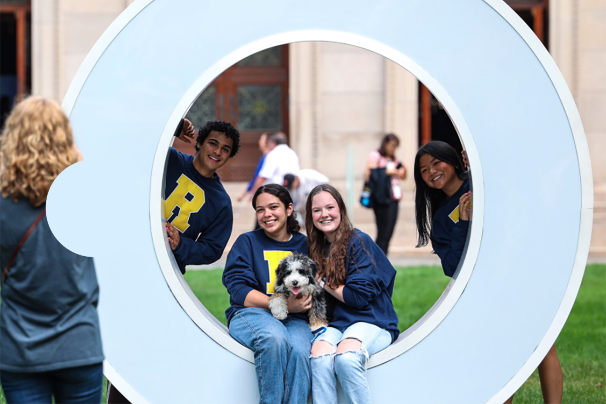 Four University of Rochester students smile while posing with a dog in a giant letter "O".
