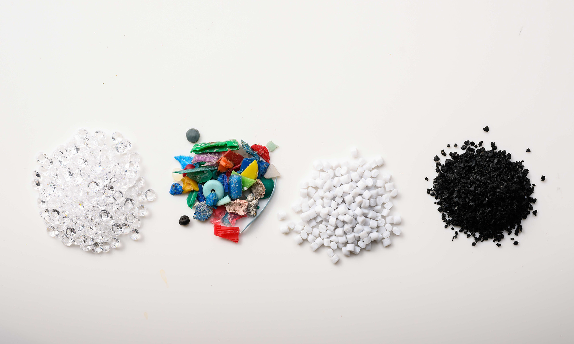 Four small groupings of plastics—one with clear plastic nurdles, one with plastic shards of many colors, one with white pellets, and one with black plastic beads—against a white background.