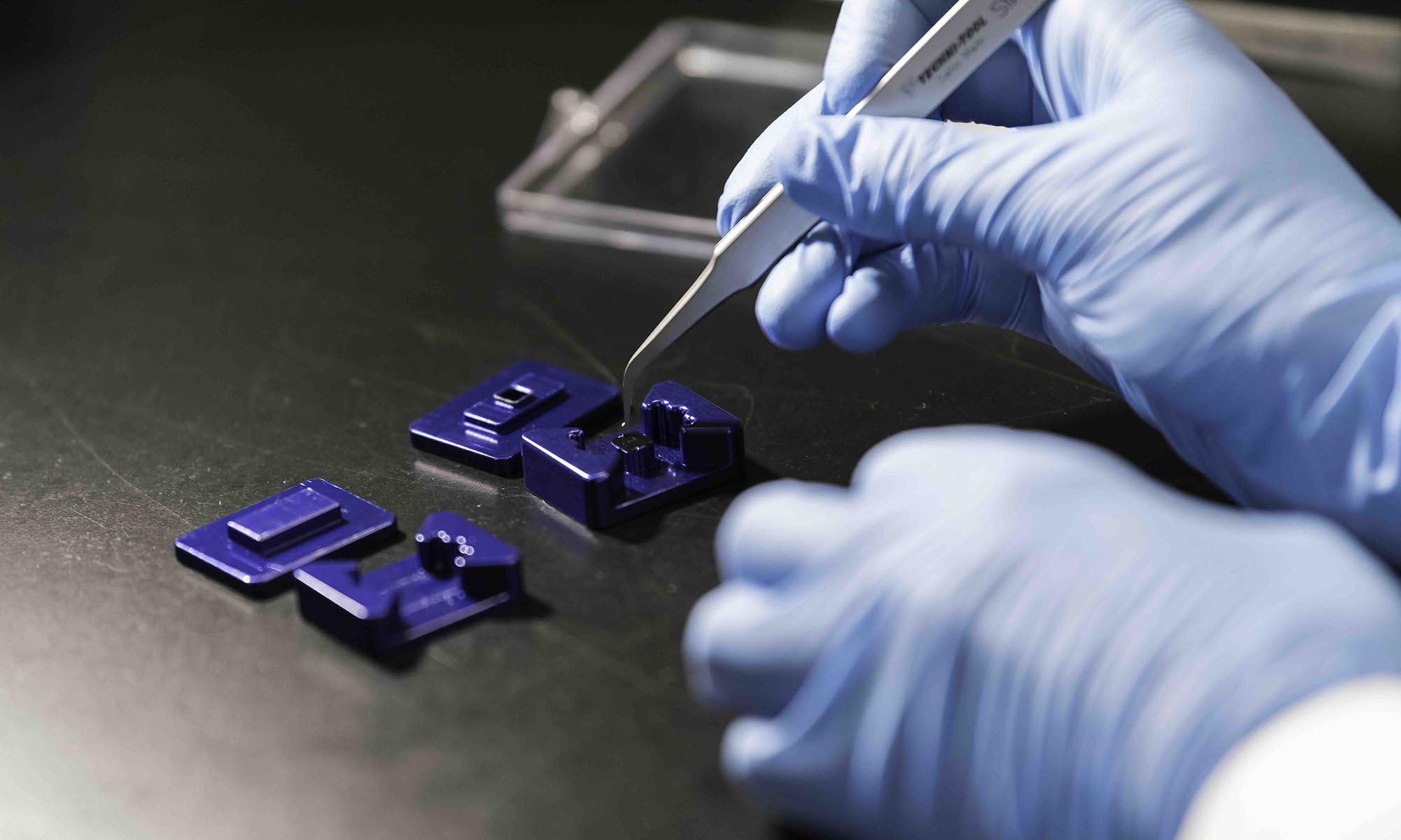 Close up of two gloved hands holding a tweezer-like tool to assemble a the components of a tissue chip platform.