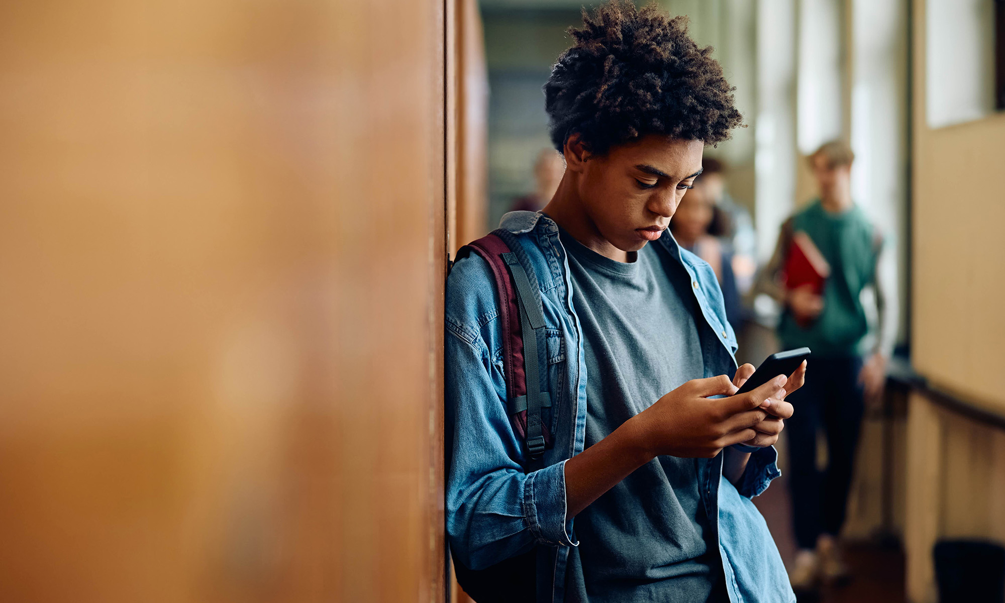 Teenager in a high school hallway leaning against a wall and looking at misinformation on his smartphone with other students in the background.