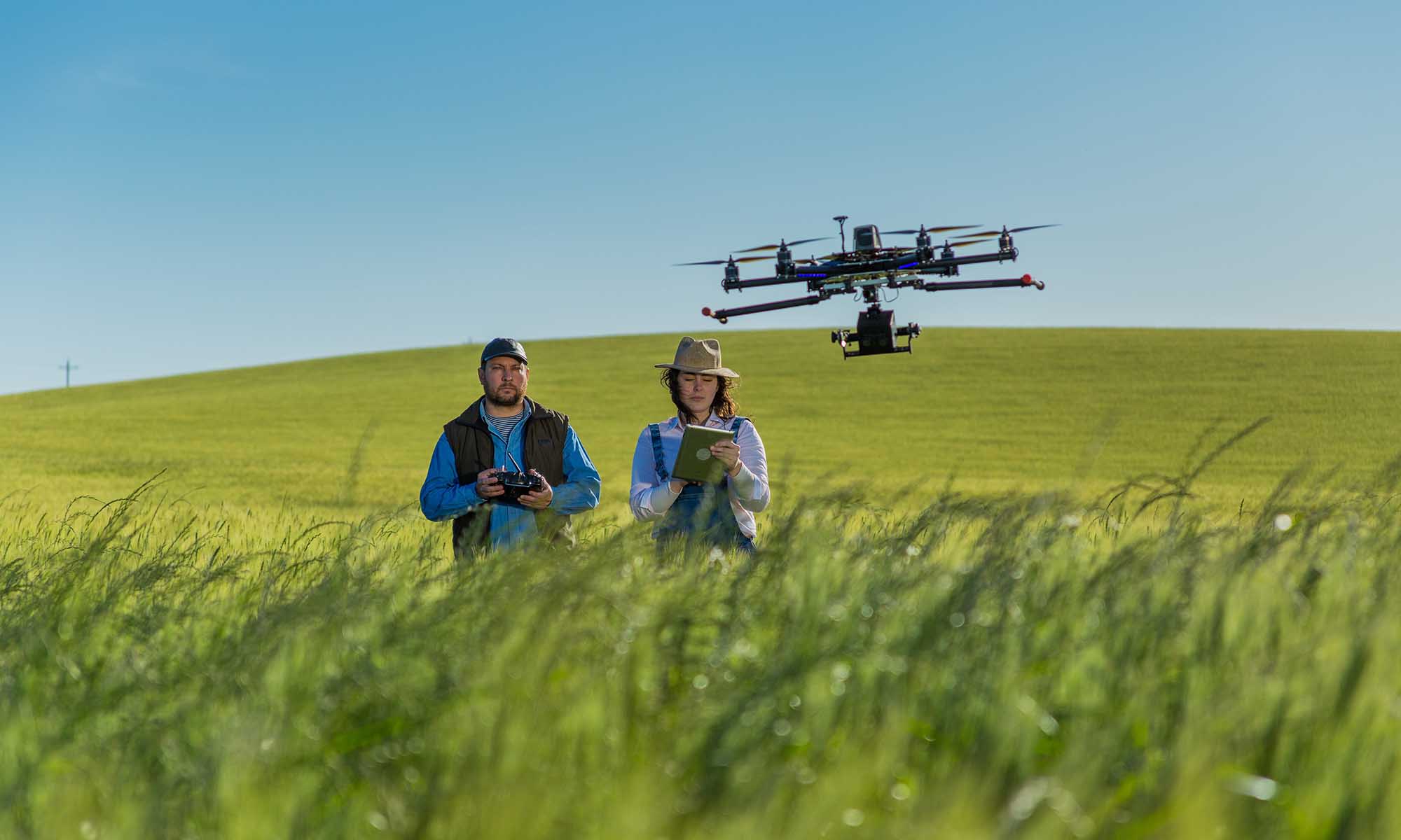 Two people in a green wheat field with a drone flying nearby overhead.