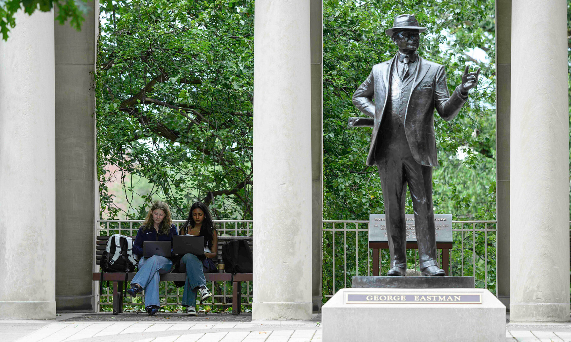 Two students sit on a bench near the George Eastman statue.