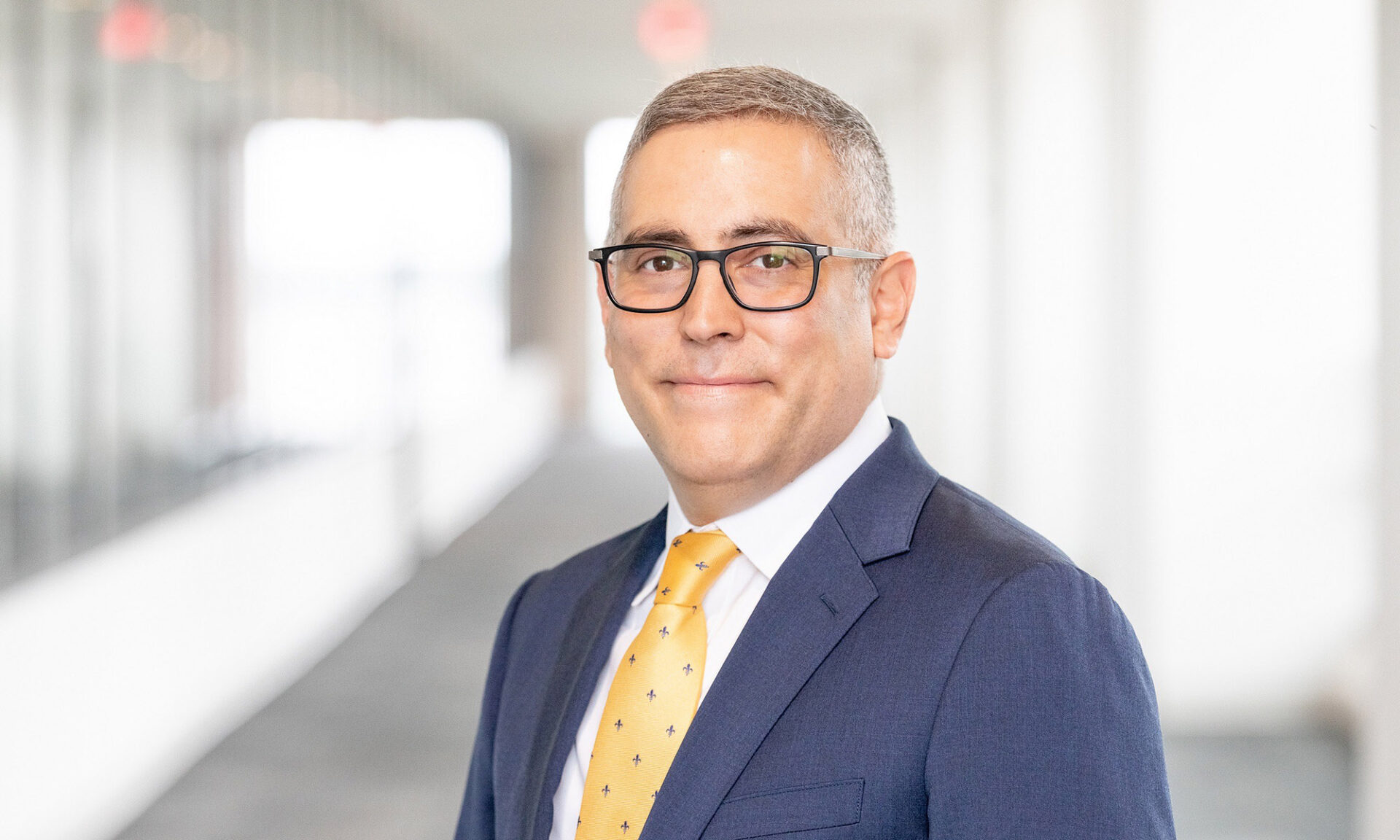 Portrait of Paul Sotoudeh in a navy suit, yellow patterned tie, and glasses, smiling in a bright hallway.