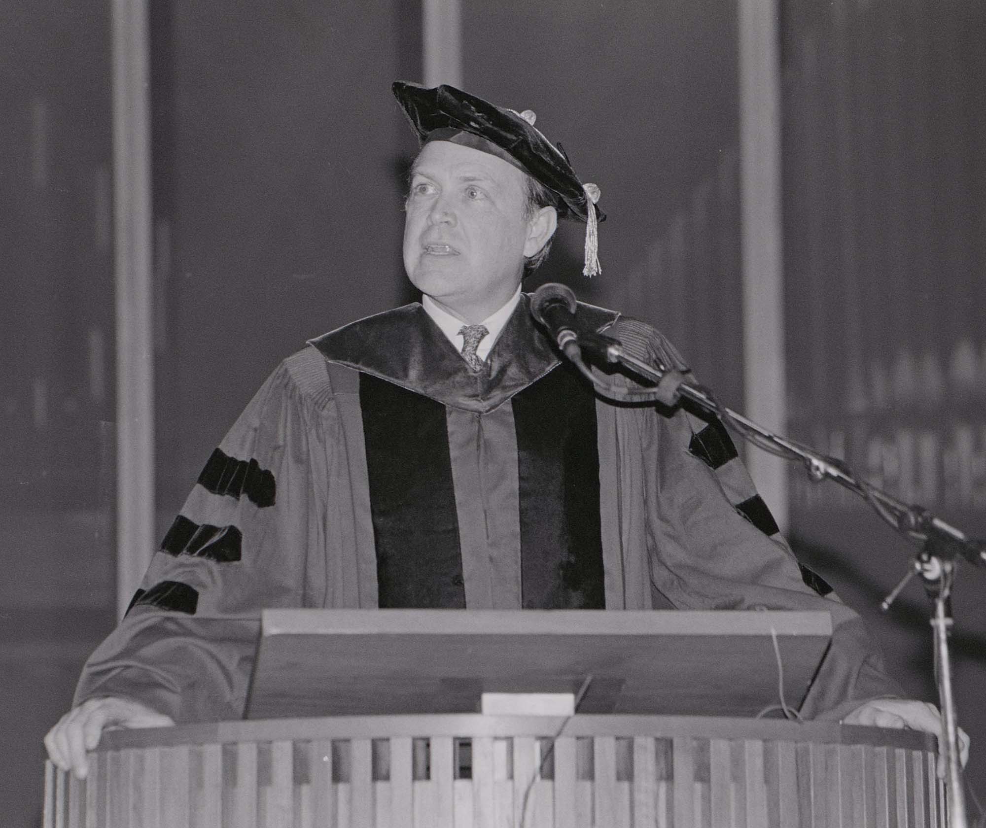 Black and white archival image of Charles Plosser in regalia speaking at a podium during his investiture as dean of the Simon Business School. 