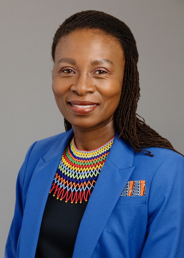 Portrait of a woman in a bright blue blazer with a colorful beaded necklace, smiling against a neutral gray background.