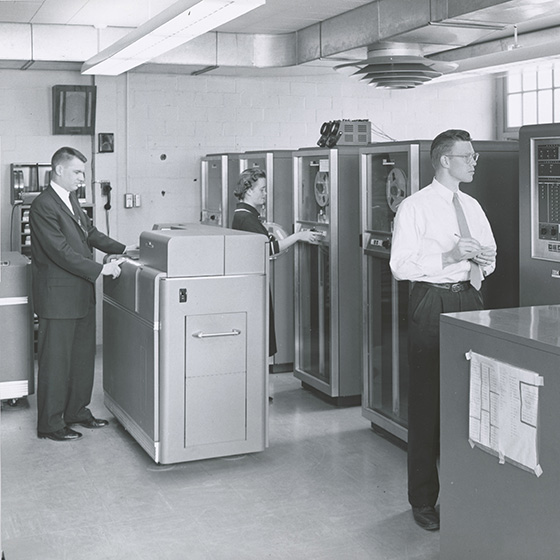 Black and white archival photo of two men and a woman in a University of Rochester computing center with tape drives, punch card reader, and other equipment.