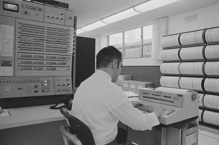 Black and white archival image of a man seen from behind working on a classical computer in the URochester computing center.