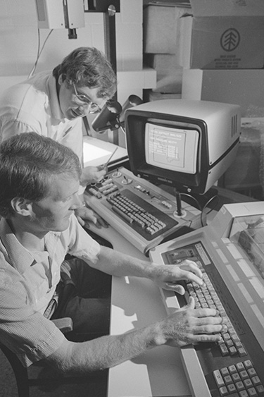 Black and white archival photo of Brian McIntyre and John Wilkerson working at a computer in 1987.