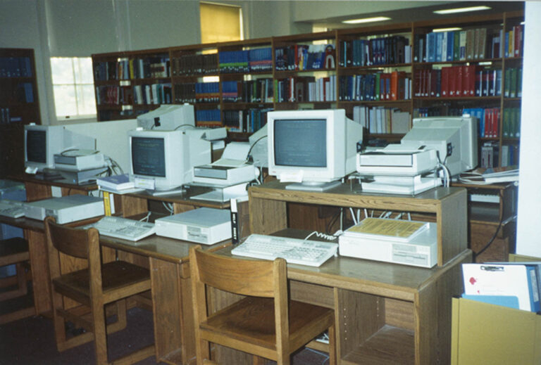 Computer terminals on desks with books and bookshelves in the background.