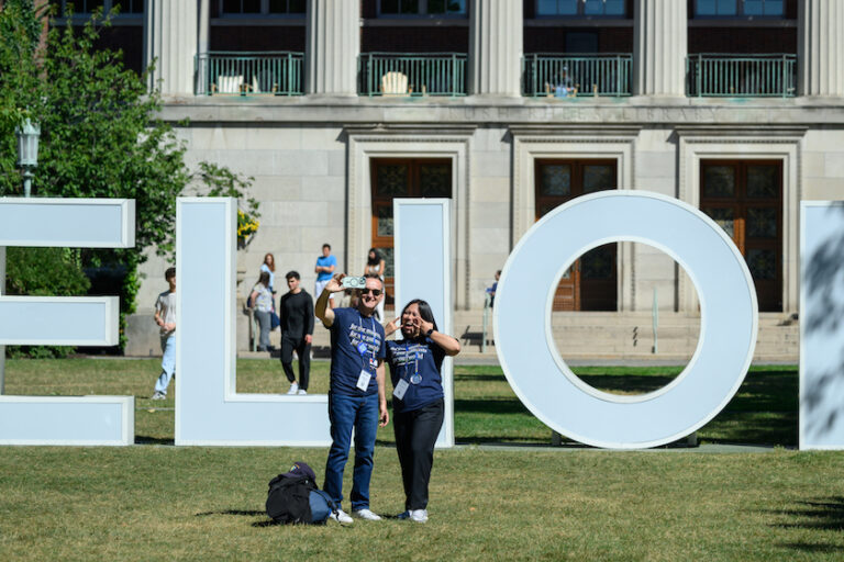 Two people take a selfie in front of the Meliora letters.