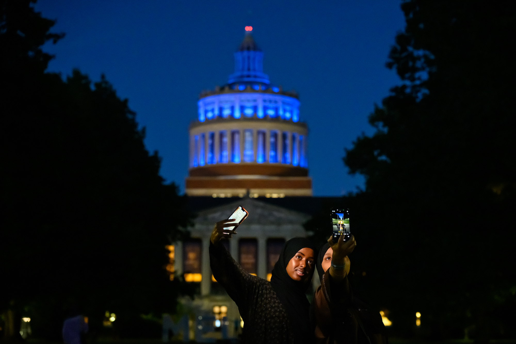 Aminah Muhumed left and Nima Mohamud (both ‘27) take selfies in front of a Rush Rhees Library lit blue for the occasion of Meliora Weekend.