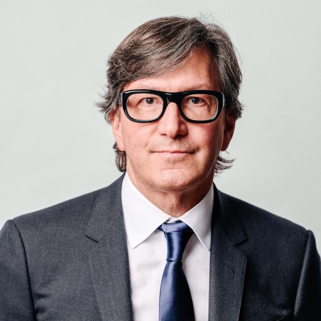 headshot of a man wearing a gray suit, blue tie, and black glasses.