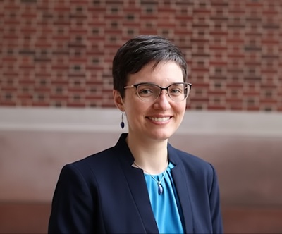 Person wearing glasses and a navy blazer over a blue blouse, smiling in front of a brick wall.