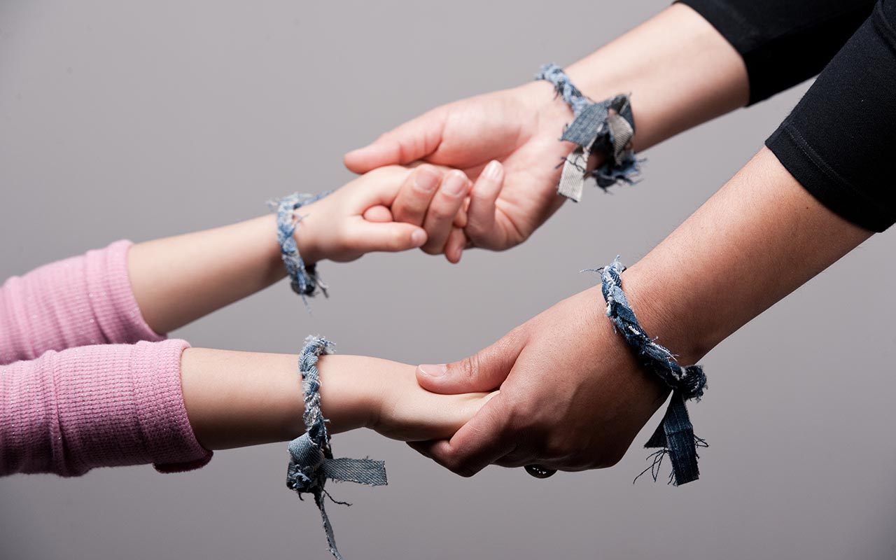 Close-up of a mother's hands holding her daughter's hands while they both wear denim bracelets. 