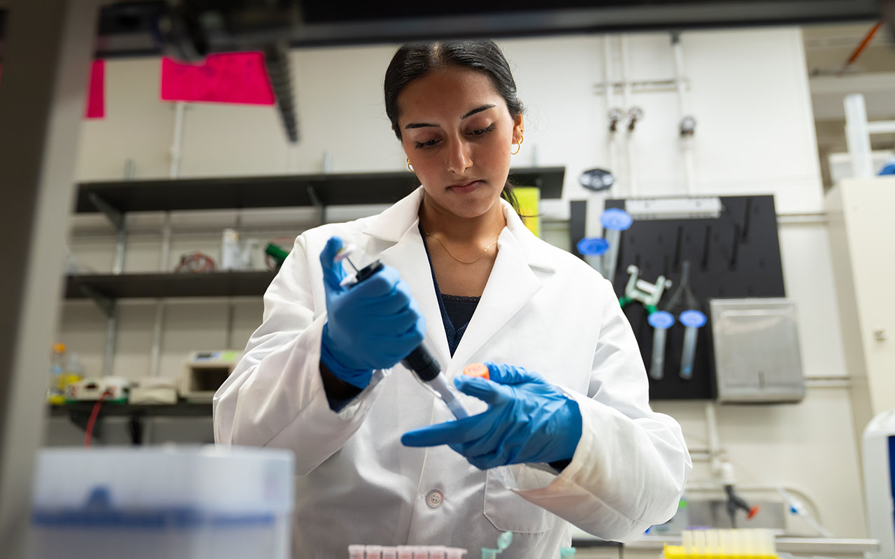 Maria Ahmed in a lab coat and safety gear empties a pipette.