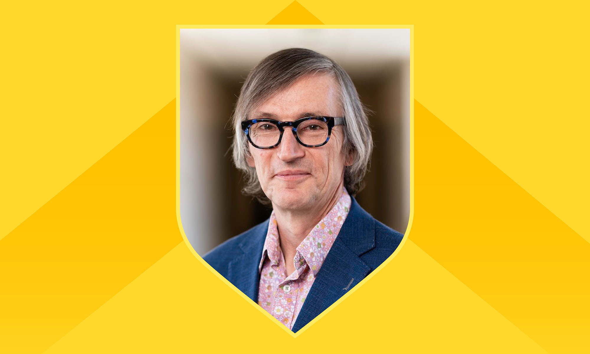 Headshot of Steve Dewhurst in a shield frame against a light yellow background with a darker yellow chevron.