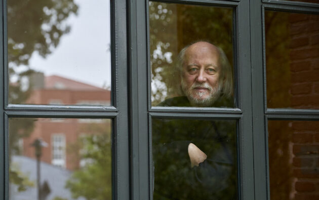 László Krasznahorkai photographed looking outside with his arms crossed through paned windows.