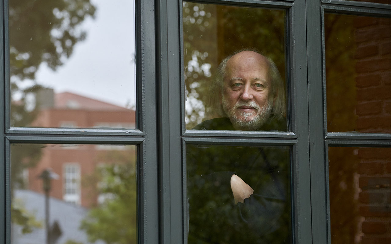 László Krasznahorkai photographed looking outside with his arms crossed through paned windows.