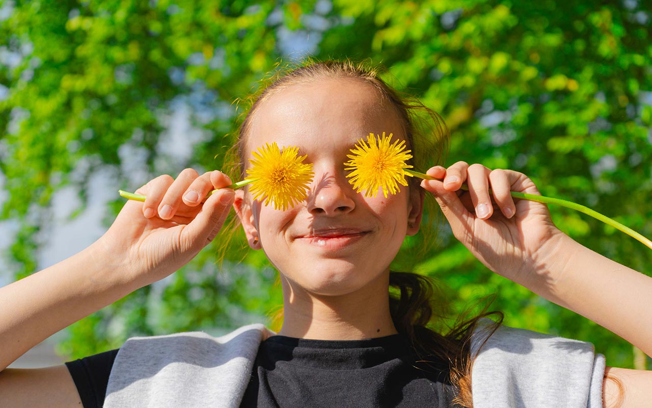 Close-up of a girl smiling and holding two dandelions in front of her eyes.