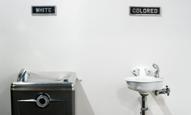 Archival photo of two water fountains next to each other. The larger one has a plaque above it reading "white" and the smaller fountain has a sign that reads "colored," illustrating American illiberalism.