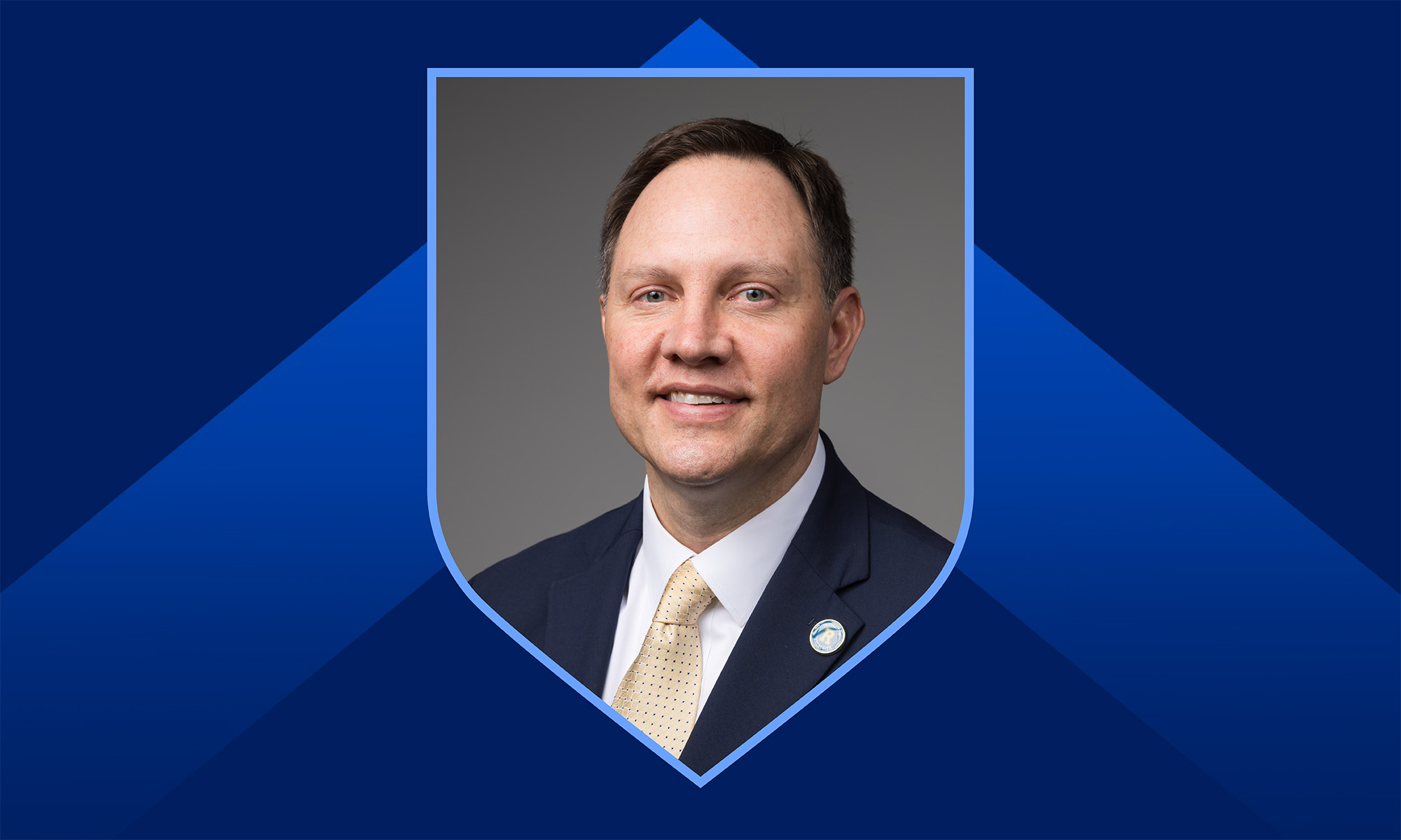 Headshot of Joshua Farrelman in a shield frame against a dark blue background with a light blue chevron.