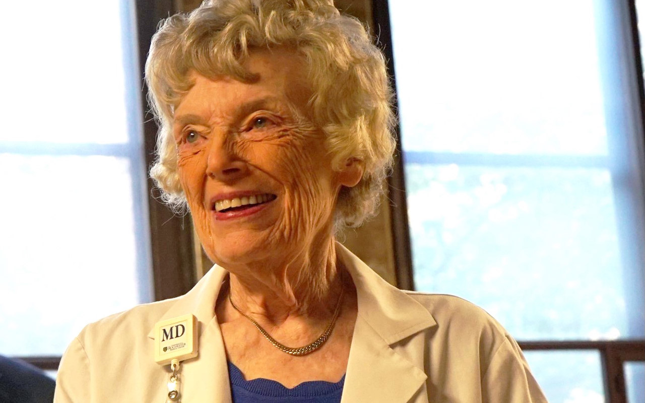 Ruth Lawrence looking off camera and smiling in a white coat with an MD badge on the collar.