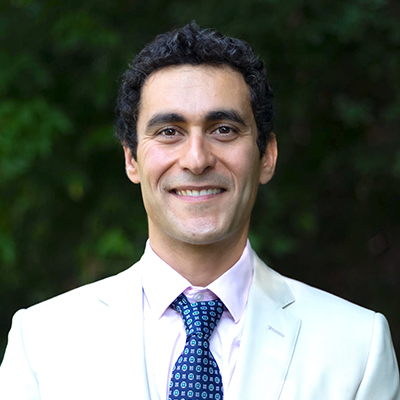 Person smiling in a light-colored suit with a patterned blue tie, standing outdoors with greenery in the background.