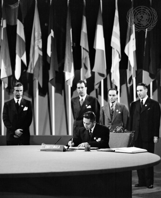 Black and white archival photo of four men in suits with flags behind them looking on as a representative from Guatemala signs a document.