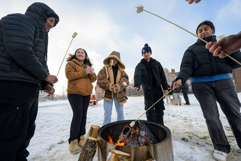 Five URochester students gather around a fire pit and make smores.