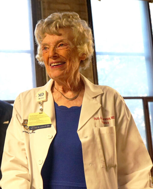 Vertical portrait of Ruth Lawrence looking off camera and smiling in a white coat with an MD badge on the collar.