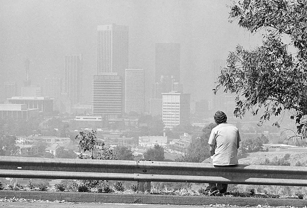 A man sits on a bench, gazing at a sprawling cityscape under a hazy sky.