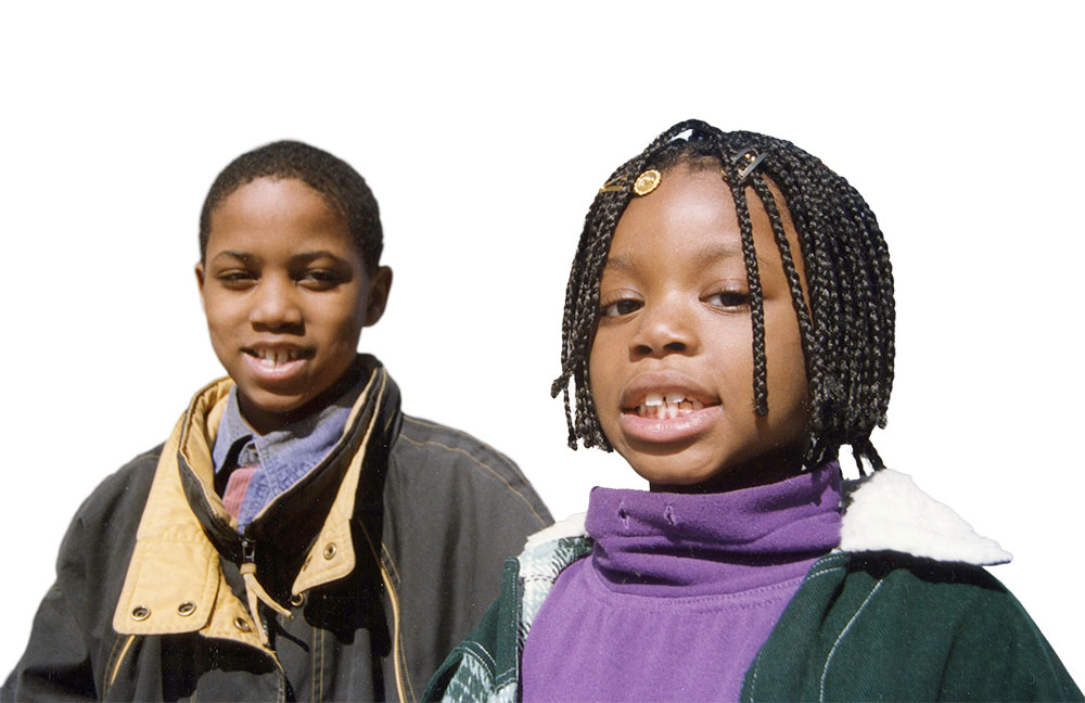 Two young children standing side by side, smiling and looking at the camera in a bright outdoor setting.
