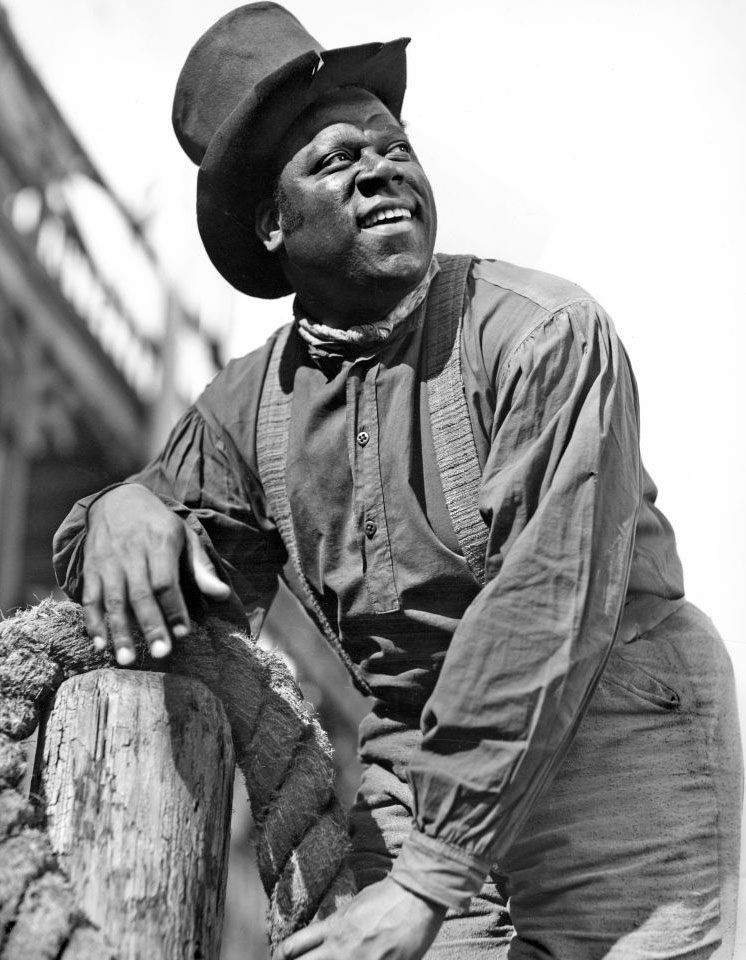 A black man in overalls and a hat leans against a wooden post, smiling softly in a rural setting.