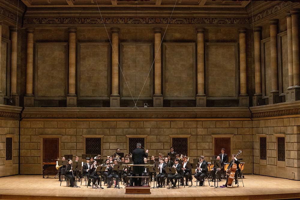 A large orchestra performs in a spacious auditorium, led by a conductor at the front.