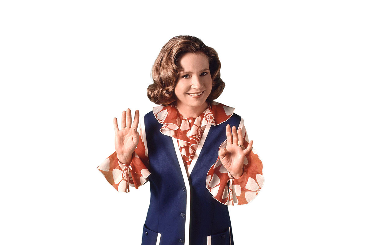A woman in a blue and orange suit and vest is smiling and making a peace sign with her right hand.